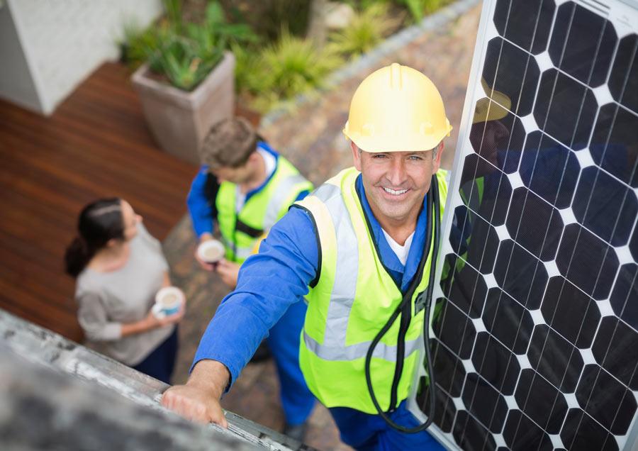 Worker in hard hat carrying a solar panel up a ladder, with two people below