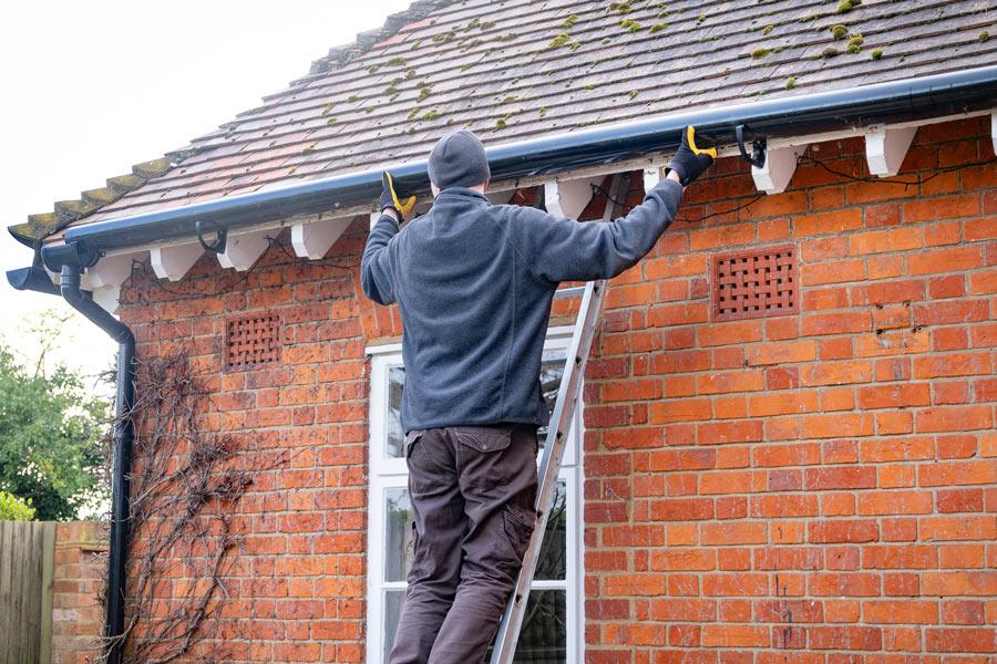 Worker on a ladder installing black guttering along the roofline of a red brick house